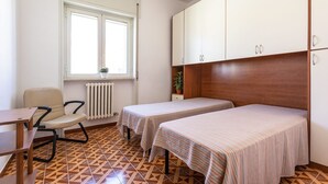 Ein kleines, sauberes Zimmer mit zwei Einzelbetten, einem Sessel, einem Tisch und einem Fenster mit Heizkörper darunter.