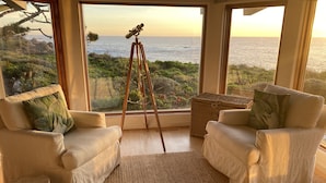 A room with two white armchairs, a telescope on a tripod, and a view of the ocean.