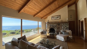 A spacious living room with a stone fireplace, large windows overlooking the ocean, and a high wooden ceiling.