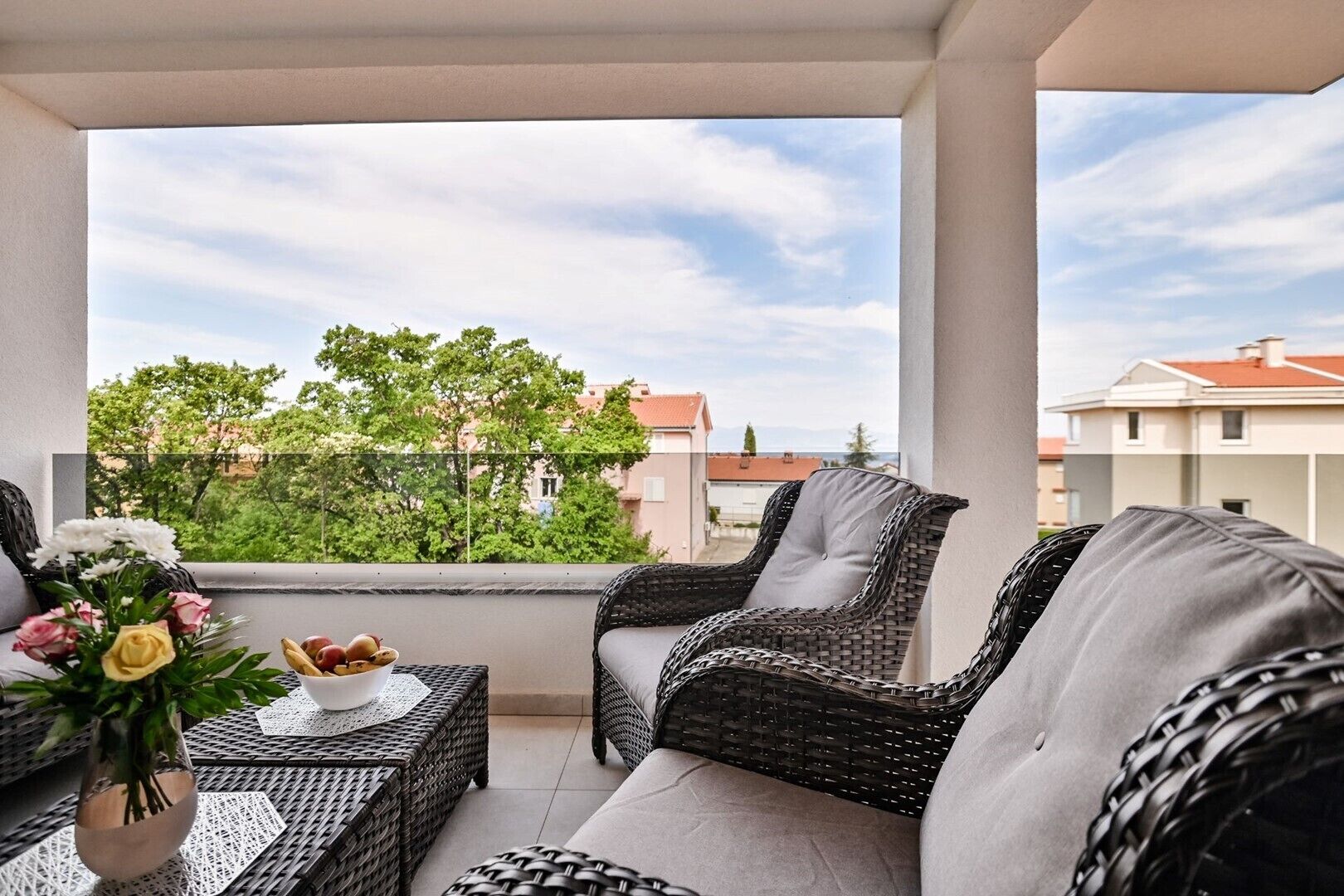 Ein Balkon mit Korbmöbeln, einem Tisch mit Blumen und Blick auf Gebäude und Bäume.