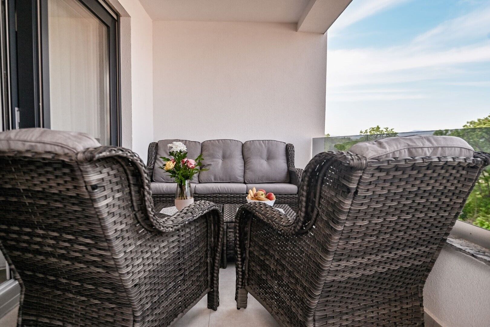 Un balcon agrémenté de meubles en osier, d’un canapé gris et d’un vase rempli de fleurs.