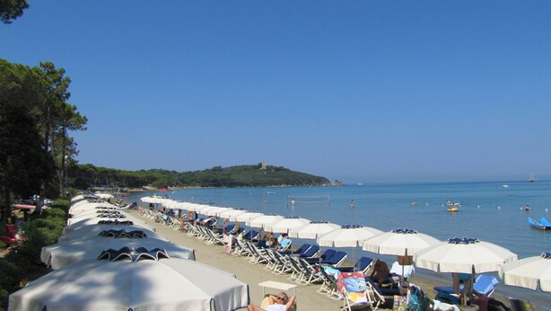 A beach with rows of sunbeds and umbrellas, a clear sky, and a distant hill with a tower. - Villa de Cuatro Familias con Jardín en el Corazón del Bosque de Pinos (Punta Ala (GR))