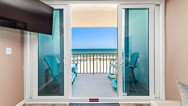 A balcony with a view of the beach, a TV mounted above, and a 'Watch Your Step' mat at the entrance. - Stunning Oceanfront Views w/Pool & Beach Access (Gulf Shores)