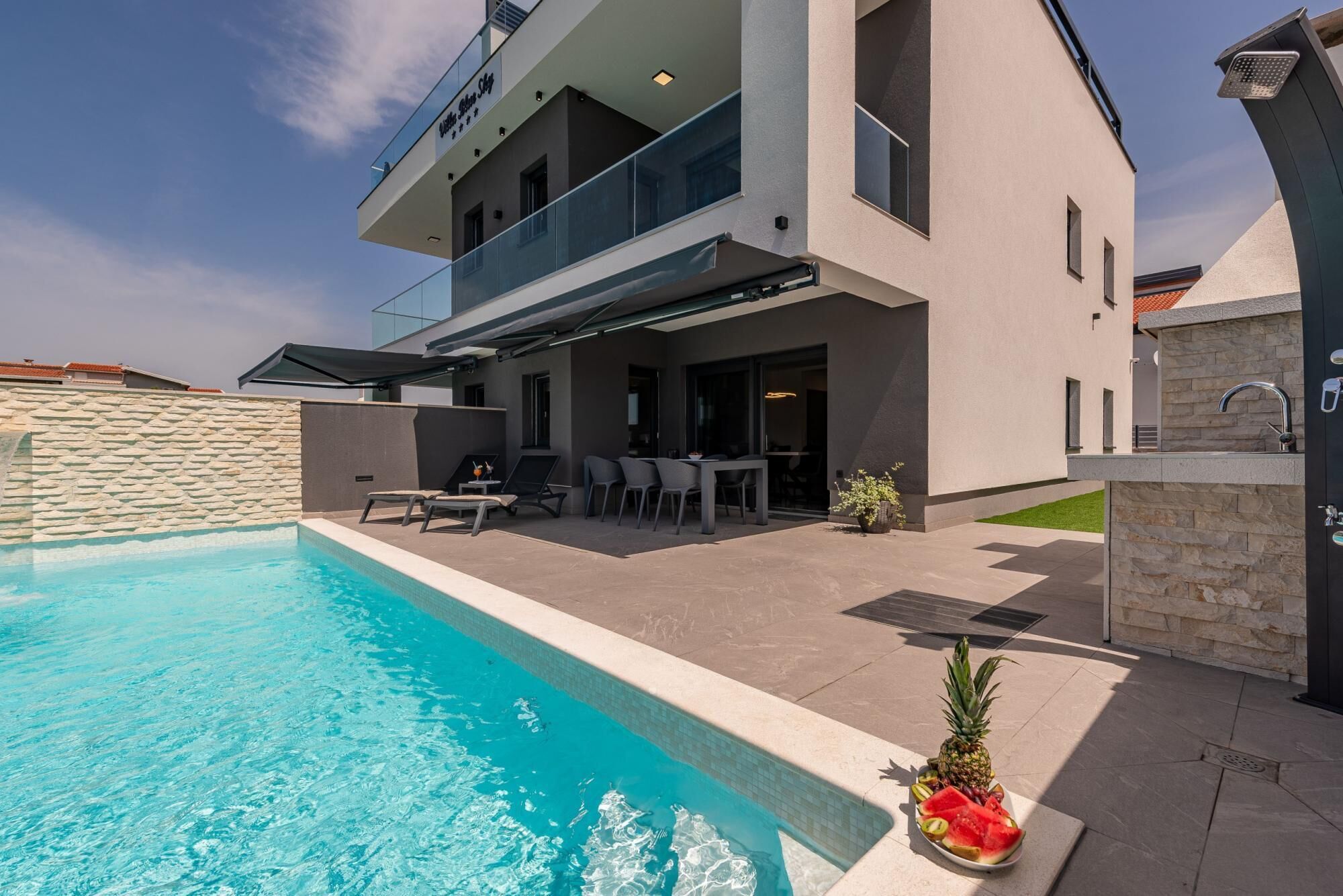 A modern two-story house with a swimming pool, outdoor seating, and a stone wall.