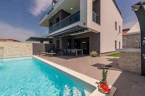 A modern two-story house with a swimming pool, outdoor seating, and a stone wall.
