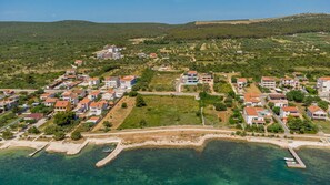 An aerial view of a coastal residential area with houses, clear blue waters, and a hilly landscape in the background.