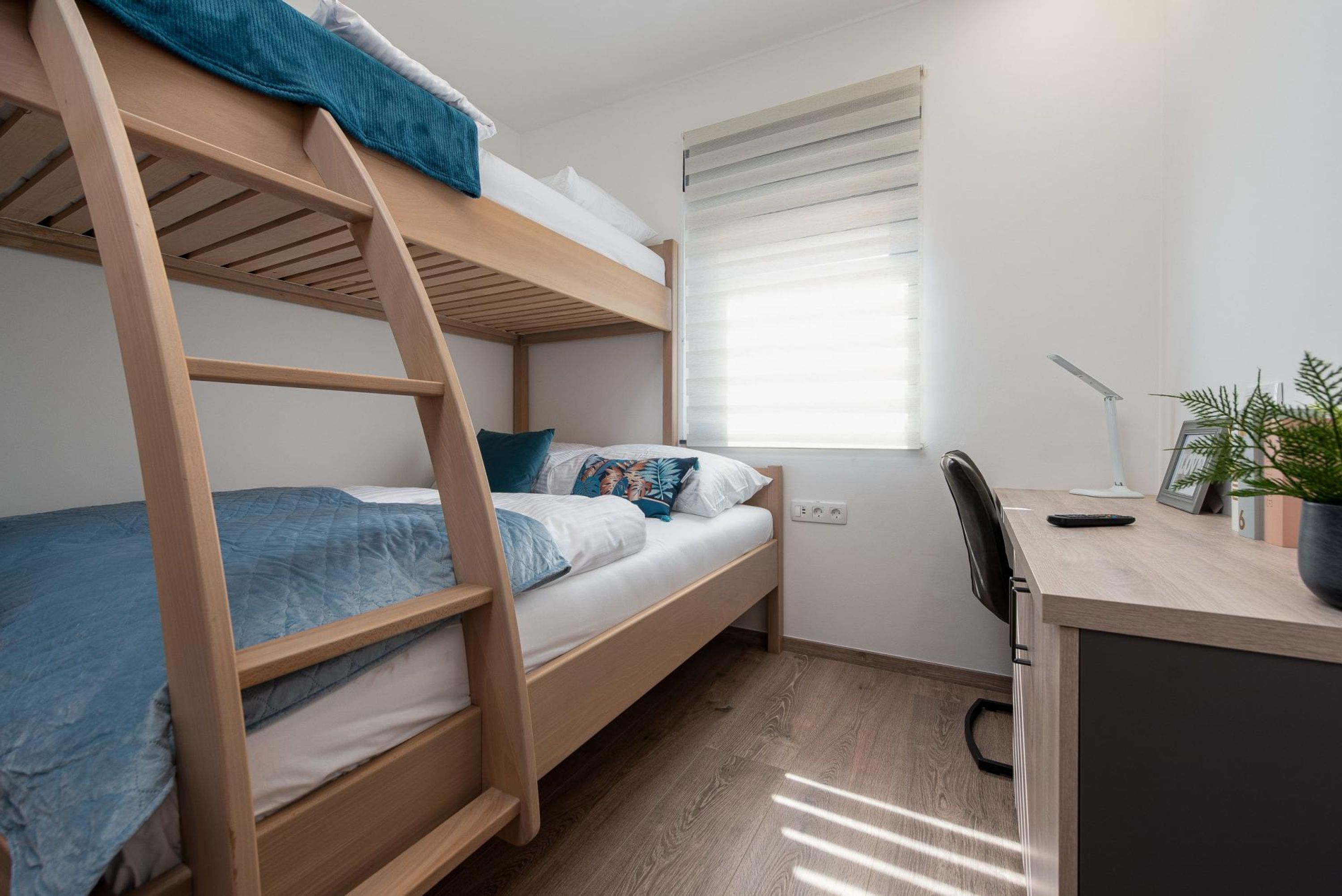 A bunk bed room with a desk and chair, a window with blinds, and a potted plant.