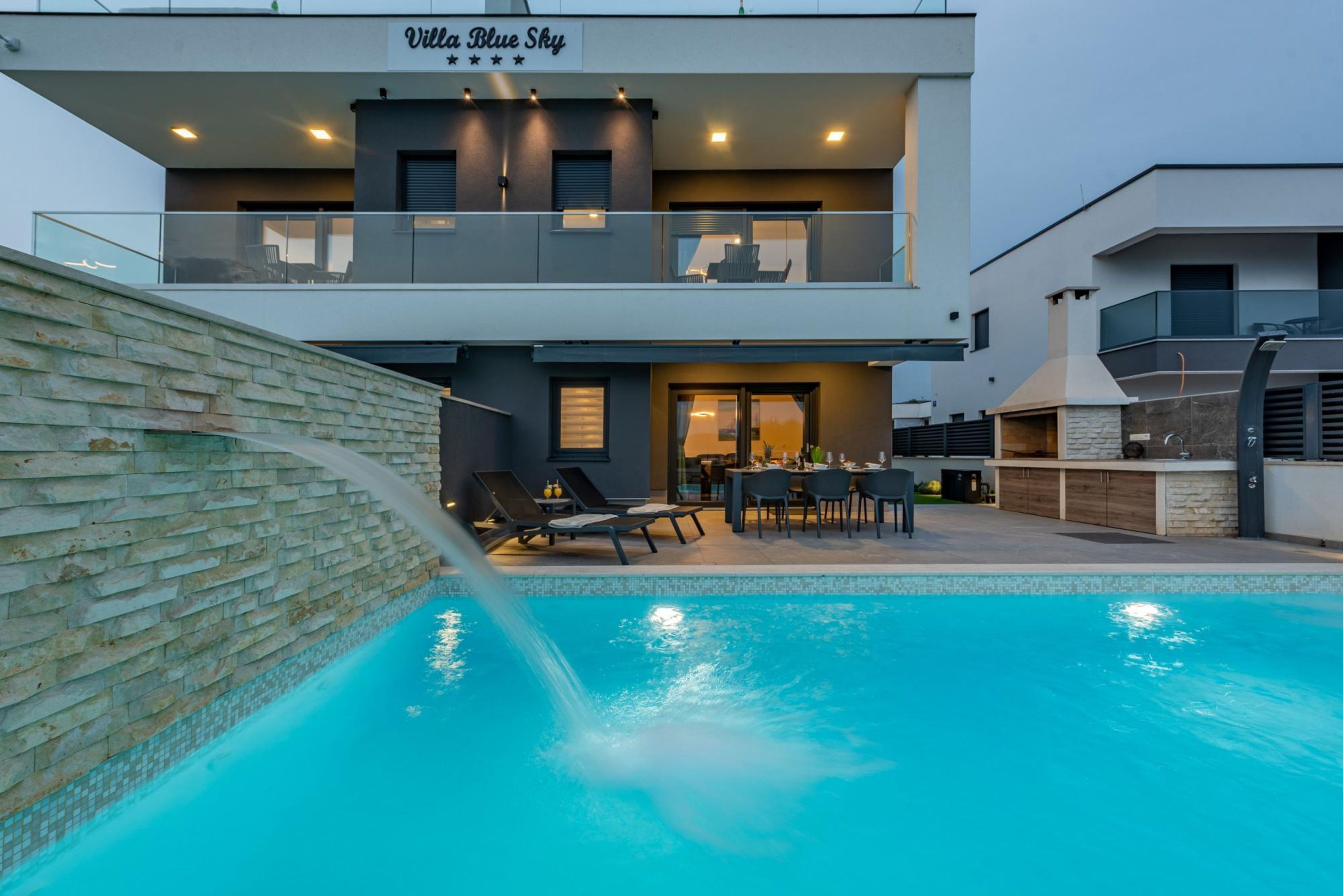 Una vila moderna de dos pisos amb piscina, mobiliari exterior i un mur de pedra.