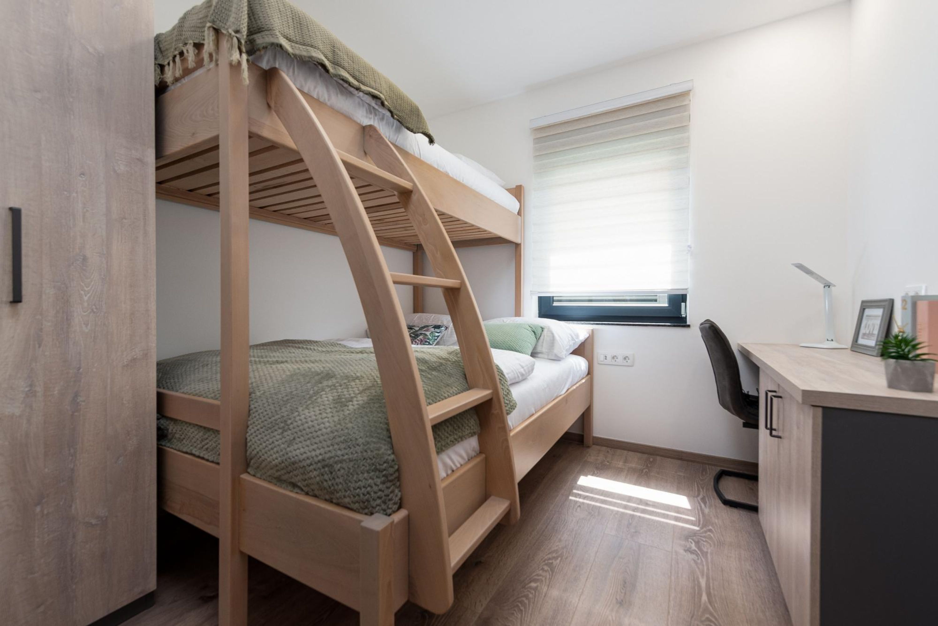 Un letto a castello con una scrivania e una sedia, una finestra con tende e una piccola pianta sulla scrivania.