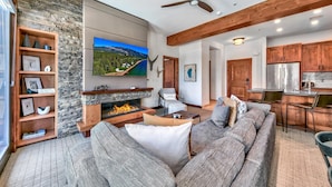 A modern living room with a stone fireplace, a large flat-screen TV, a sofa, and a dining area with wooden cabinets and chairs.