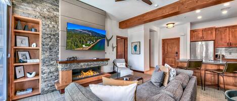 A modern living room with a stone fireplace, a large flat-screen TV, a sofa, and a dining area with wooden cabinets and chairs.