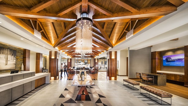 A spacious lobby with a high ceiling, wooden beams, a central chandelier, a reception desk, seating areas, and a large TV screen.