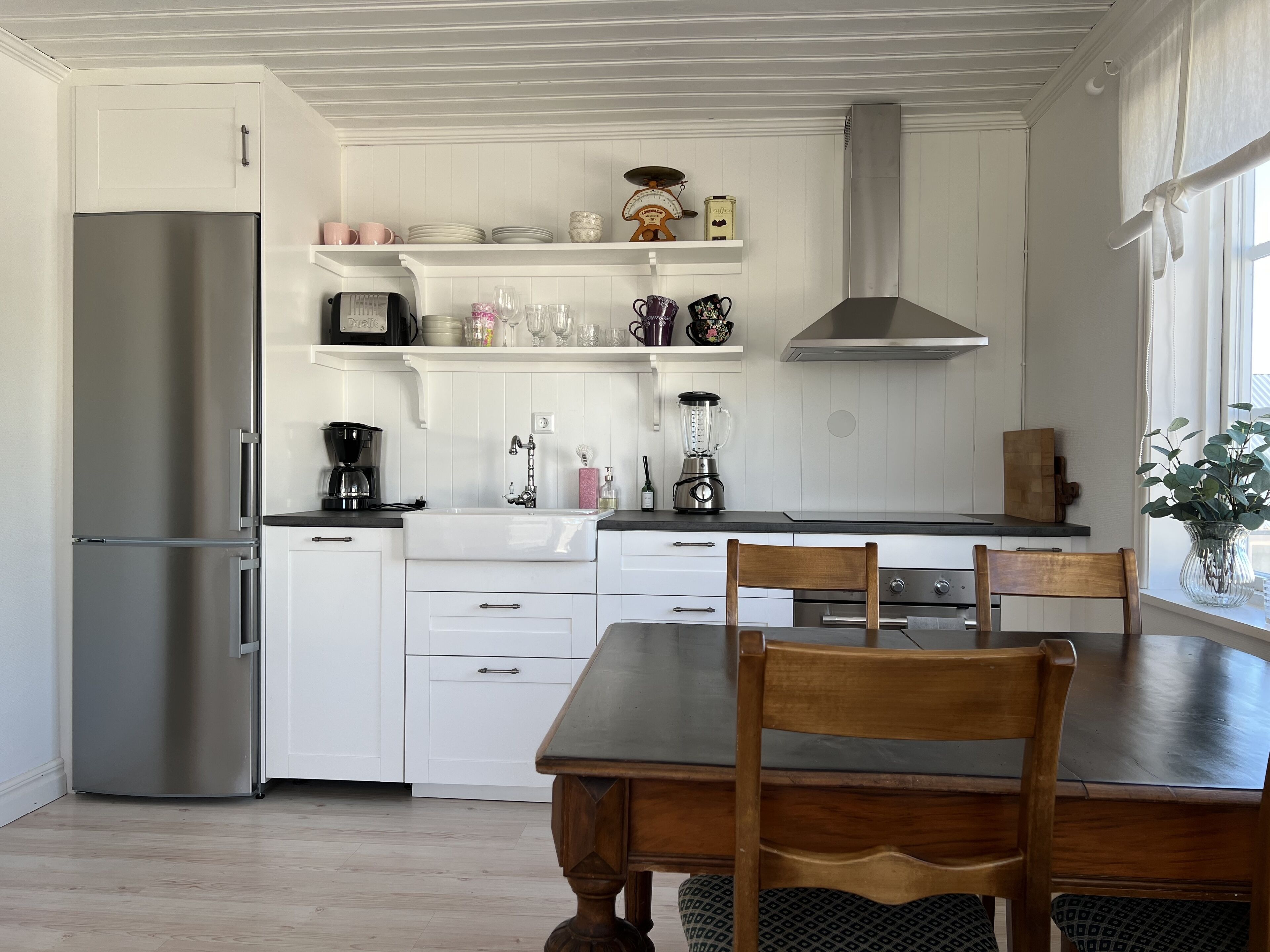 A modern kitchen with white cabinetry, a stainless steel refrigerator, a wooden dining table with chairs, and a window with a view.