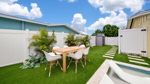 A backyard with a wooden table and white chairs, surrounded by artificial grass and plants. - Mykonos: Beautifully updated with heated pool, moments to the Gulf (Anna Maria)
