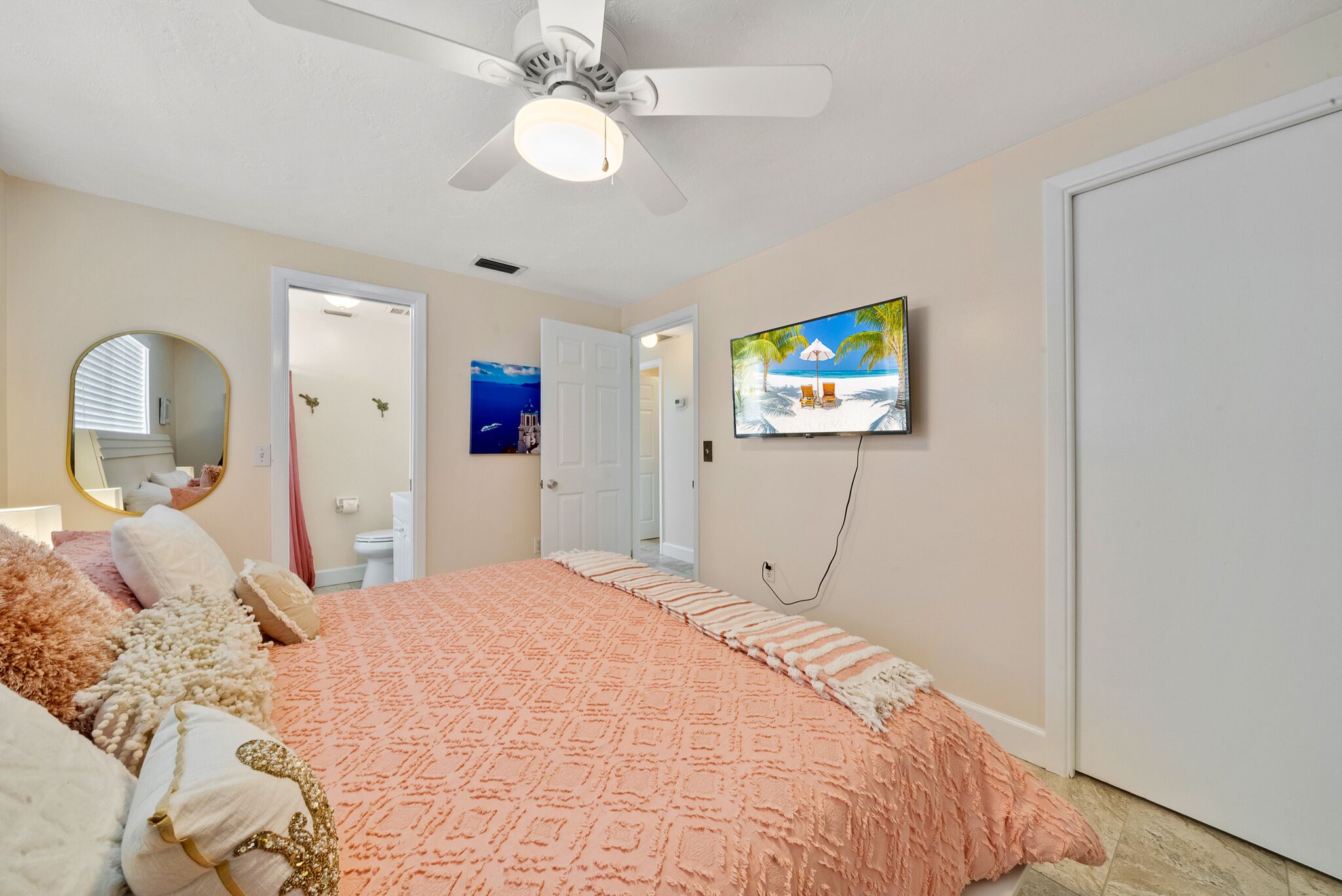 A bedroom with a large bed, a ceiling fan, a TV on the wall, and a door leading to another room.