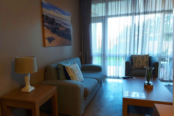 Una sala de estar con un sofá, una mesa de centro y una lámpara. Hay un cuadro en la pared y una ventana con cortinas.