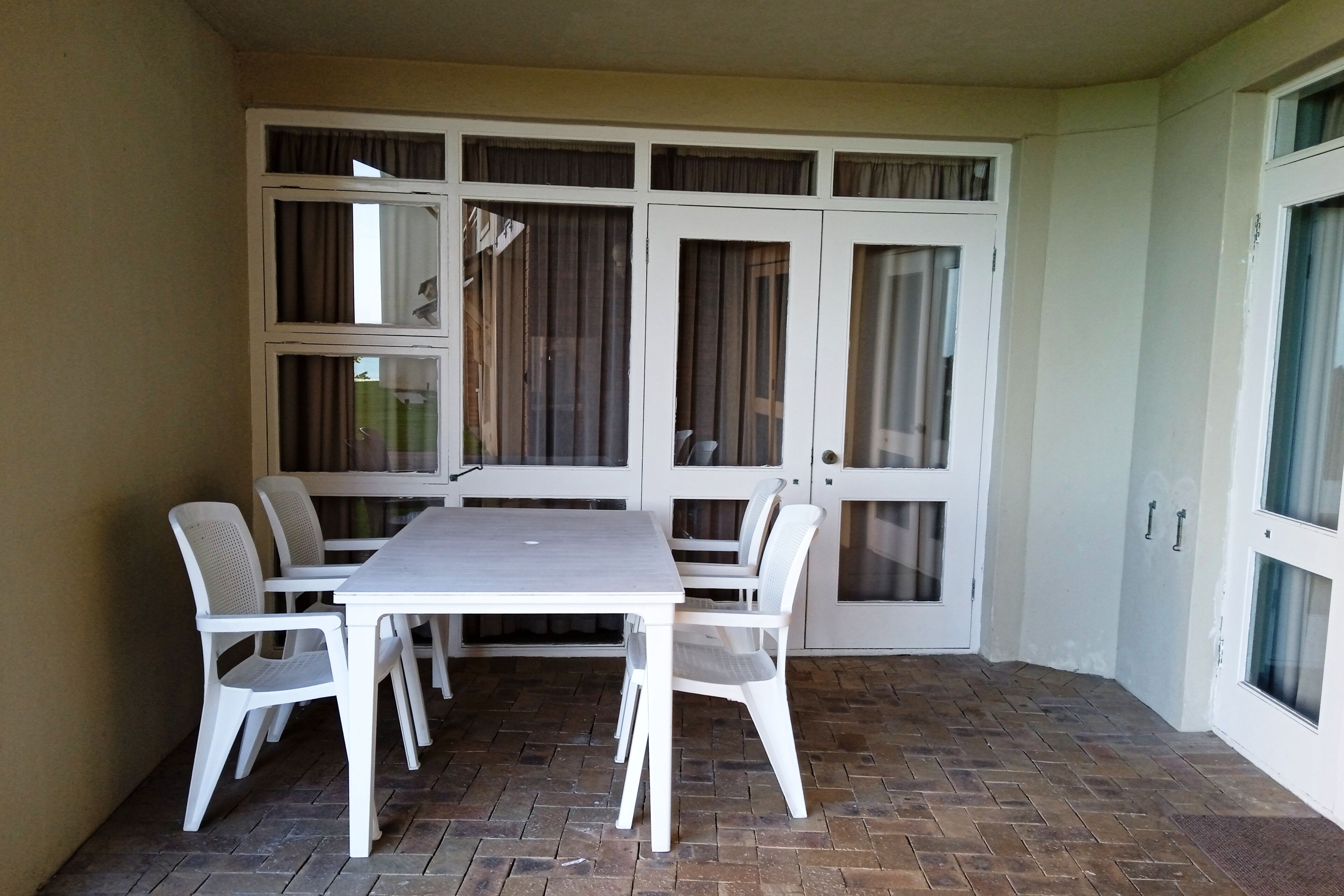 Une pièce avec une table et des chaises blanches, une grande fenêtre avec des rideaux et un sol carrelé.