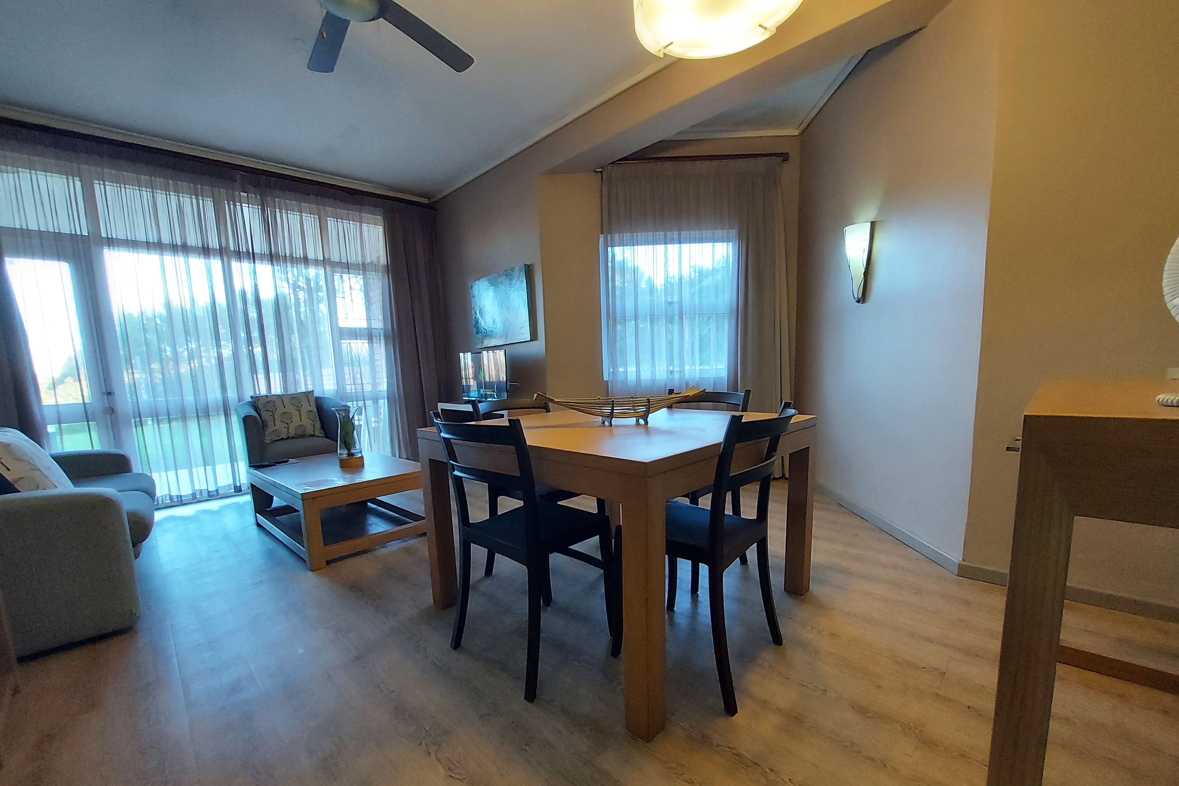 A living room with a wooden dining table, four chairs, a sofa, and a TV on the wall.