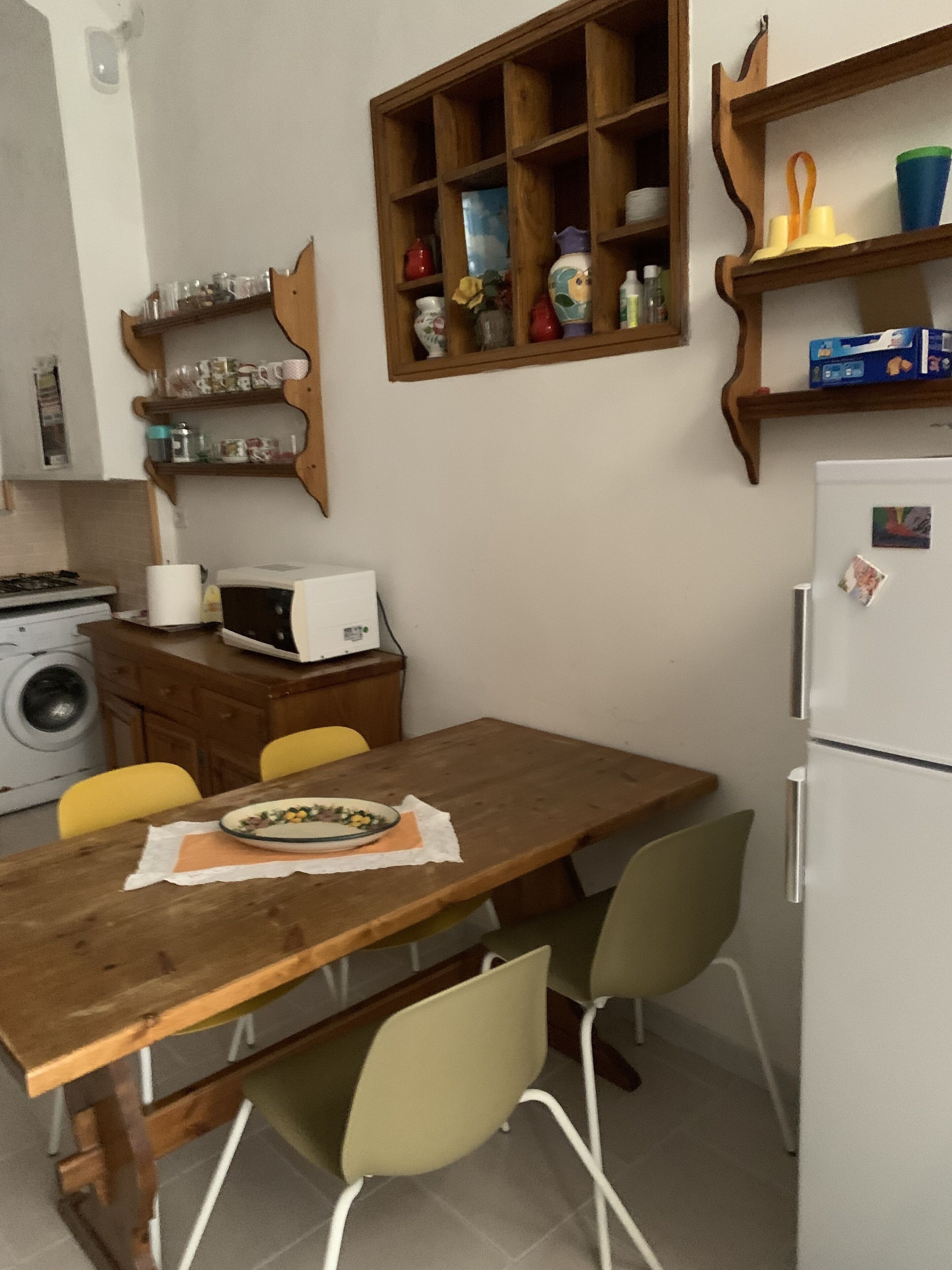 Una cucina compatta con un tavolo in legno, due sedie, un forno a microonde, una lavatrice e mensole piene di utensili da cucina.