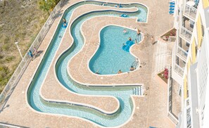 Vue aérienne d’un complexe de piscines comprenant plusieurs couloirs et une piscine centrale, le tout entouré d’une terrasse carrelée et d’un bâtiment doté de balcons.