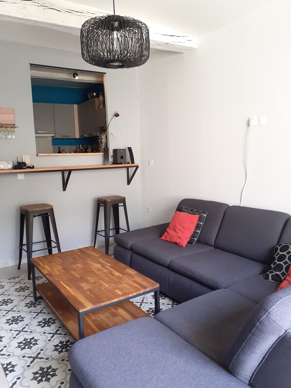A modern living room with a grey sofa, a wooden coffee table, two bar stools, a hanging light fixture, and a kitchen area with a blue backsplash visible through the mirror.