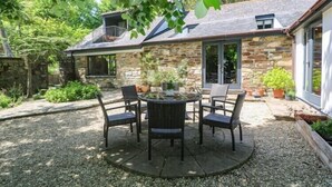 Ein Steinhaus mit einer Terrassenfläche, ausgestattet mit einem runden Tisch und Stühlen, umgeben von Topfpflanzen und einem Kiesgrund.