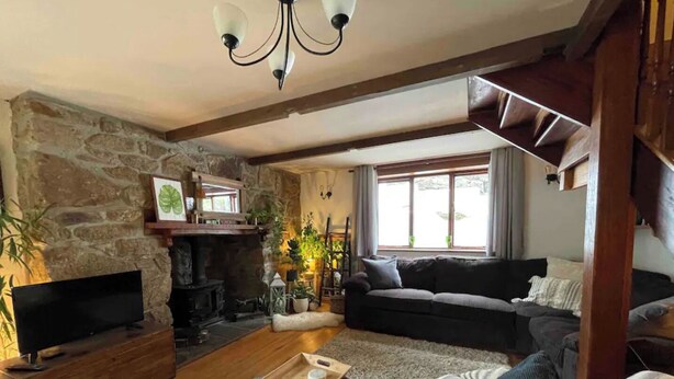 Ein gemütliches Wohnzimmer mit Steinwand, Holzbalken, Kamin, einer Couch, einem Fernseher und einem Fenster mit Vorhängen.