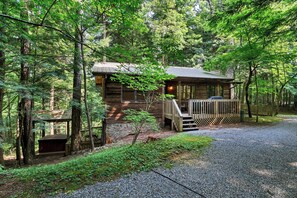 Une cabane dotée d’une terrasse en bois et de fondations en pierre, entourée d’une forêt dense.