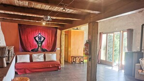 A spacious room with a wooden ceiling, a red tapestry with a meditative figure, a cushioned bench, and a large window offering a view of greenery. - Very pretty, bright, south-facing sheepfold on an organic farm in the Verdon.  (Var)