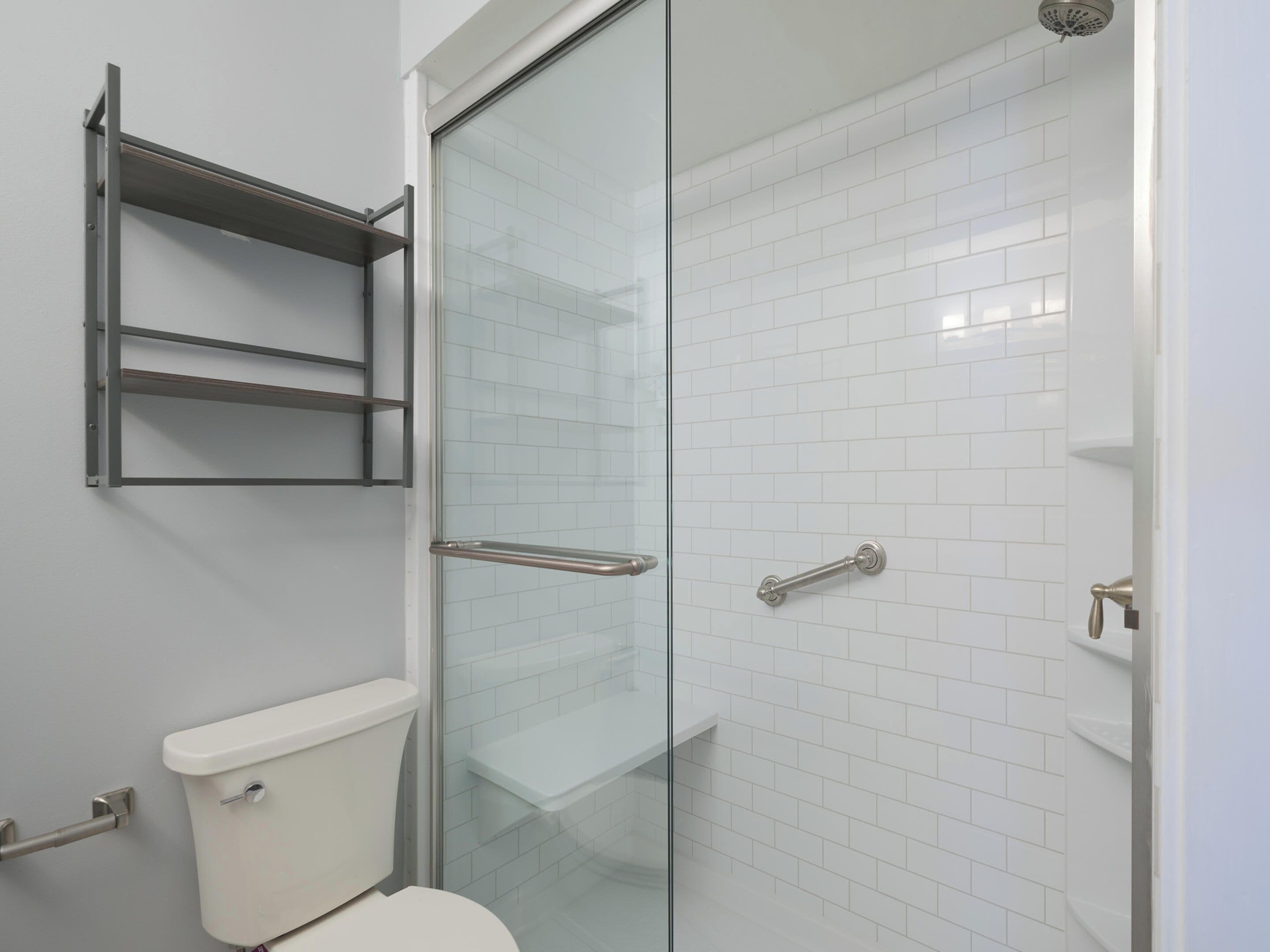 A bathroom with a glass shower enclosure, white tiled walls, and a toilet with a grab bar.