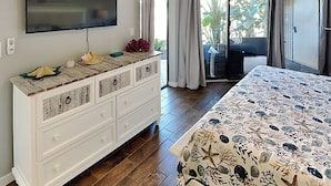 Une chambre avec un lit, un téléviseur fixé au mur, une commode et une porte-fenêtre coulissante donnant sur un balcon agrémenté de plantes.