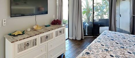 A bedroom with a bed, a TV mounted on the wall, a dresser, and a sliding glass door leading to a balcony with plants.