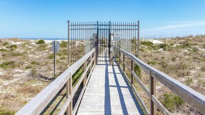 Une promenade en bois mène à une entrée fermée par un portail, d’où l’on a une vue dégagée sur la plage et l’océan.