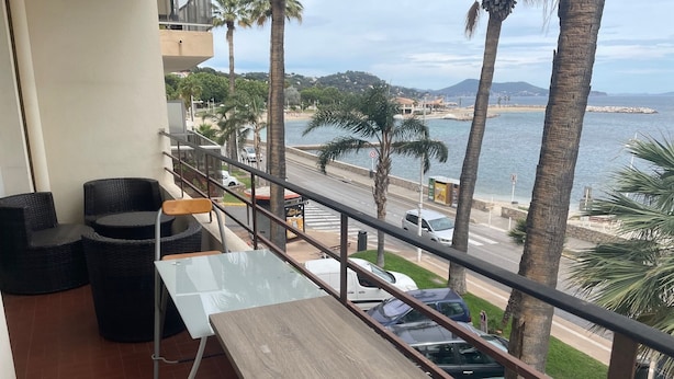 Un balcon avec des fauteuils et une table en osier, donnant sur une plage bordée de palmiers et une route où sont garées des voitures.