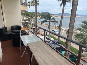 A balcony with wicker chairs and a table, overlooking a beach with palm trees and a road with parked cars. - Apartment T3 beachfront 2Mn walk (Toulon)