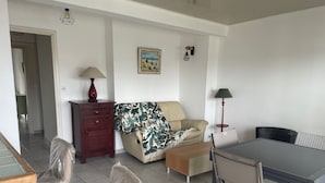 Un salon avec un canapé, une table à manger et des chaises. On y trouve une lampe, un tableau accroché au mur et un petit meuble.