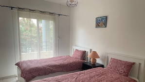 Une chambre avec deux lits, une table de chevet, une lampe, une fenêtre avec des rideaux et un tableau encadré au mur.