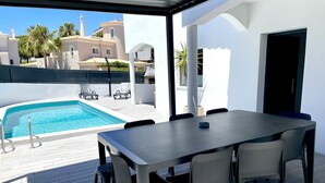 A covered outdoor area with a pool, a dining table with chairs, and a view of buildings in the background.