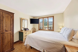 A bedroom with a bed, a wooden wardrobe, a dresser, a television, and a window with a view.