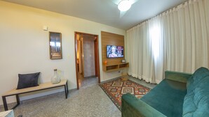Una sala de estar con un sofá verde, un banco de madera, un televisor sobre una estantería de madera, un ventilador de techo y una alfombra estampada.
