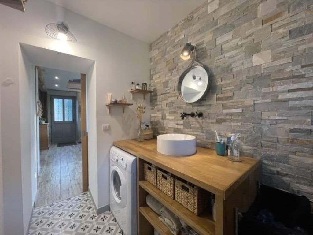 Un baño moderno con pared de piedra, un espejo redondo, un tocador de madera y una lavadora.