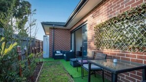 A modern outdoor patio with a glass table and chairs, a brick wall, and a green artificial turf area. - ADORABLE 2 Bedroom + Study Villa - free parking. (Rosebud)