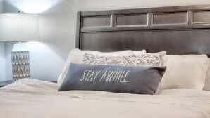 Een net opgemaakt bed met een donker kussen waarop 'VERBLIJF HIER' staat.