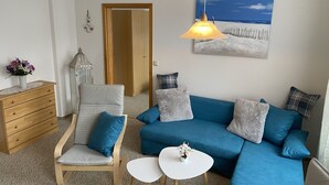 A living room with a blue sofa, a wooden chair, a small white table, a wooden dresser, a white vase with flowers, a wall-mounted lamp, and a painting of a beach scene. - Vacation at the Baltic Sea - beautiful vacation apartment for 4 persons (Warnow)