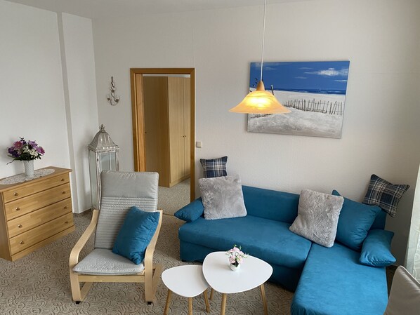 Un salón con un sofá azul, una silla de madera, una mesita blanca pequeña, un aparador de madera, un jarrón blanco con flores, una lámpara de pared y un cuadro con una playa.