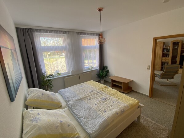 Un dormitorio con cama, dos ventanas con cortinas, un radiador, una mesita de madera, una planta en maceta y una silla.