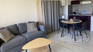 A modern living room with a grey sofa, a small wooden table, and a dining area with a round table and chairs. - Grand Studio à la Coudouliere Proche de la Plage (Six-Fours-les-Plages)
