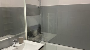 A modern bathroom with a glass shower enclosure, a white sink with a mirror, and grey tiled walls. - Grand Studio à la Coudouliere Proche de la Plage (Six-Fours-les-Plages)