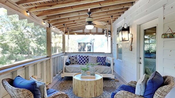 A covered porch with a wooden ceiling, a cushioned bench with patterned cushions, wicker chairs with blue cushions, and a round wooden coffee table. - The Fort | Private Pool, Fire Pit Arcade Games (Tybee Island)
