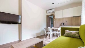 A modern living room with a green sofa, a flat-screen TV mounted on the wall, a wooden bench, and a small dining area with white chairs and a table. - Holiday apartment with terrace (Fažana)
